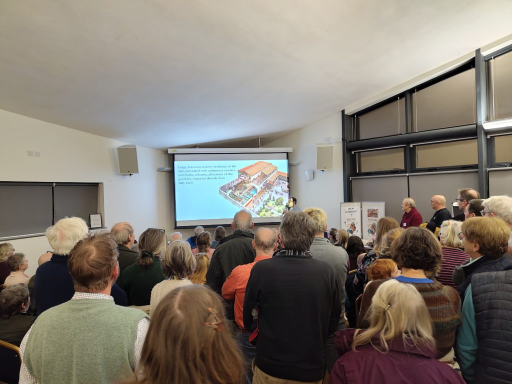 People standing and seated watching a talk. On the screen is an illustration of a Roman villa.