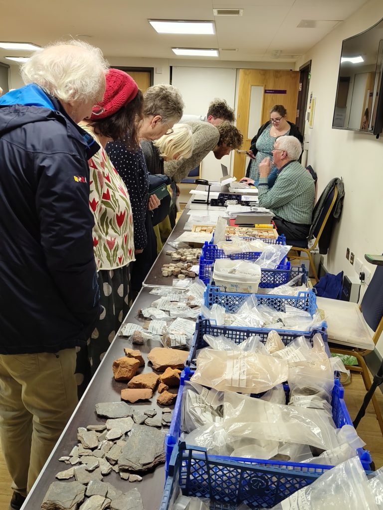A variety of finds from the villa site are spread out on the table with people looking at them. Further up the table people are taking to John about his finds.