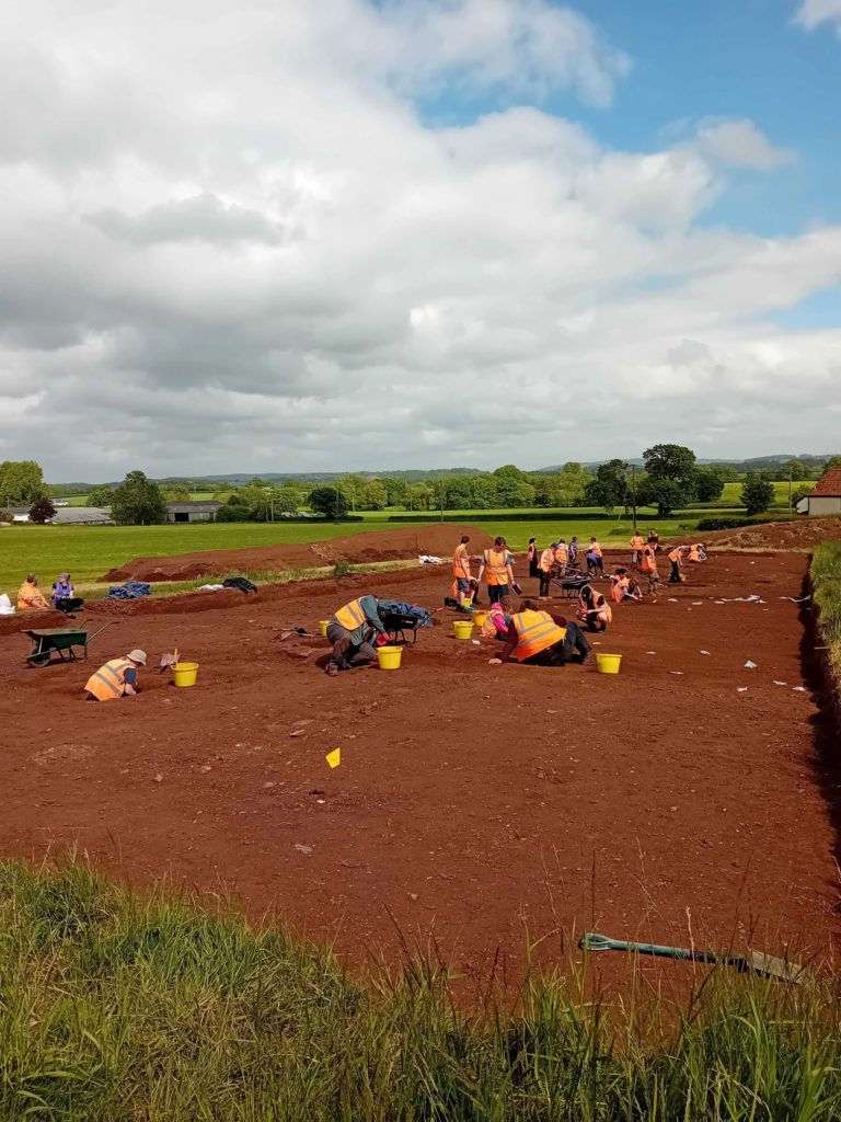 A large excavation trench, with c.20 students in high-vis orange vests excavating on the site.