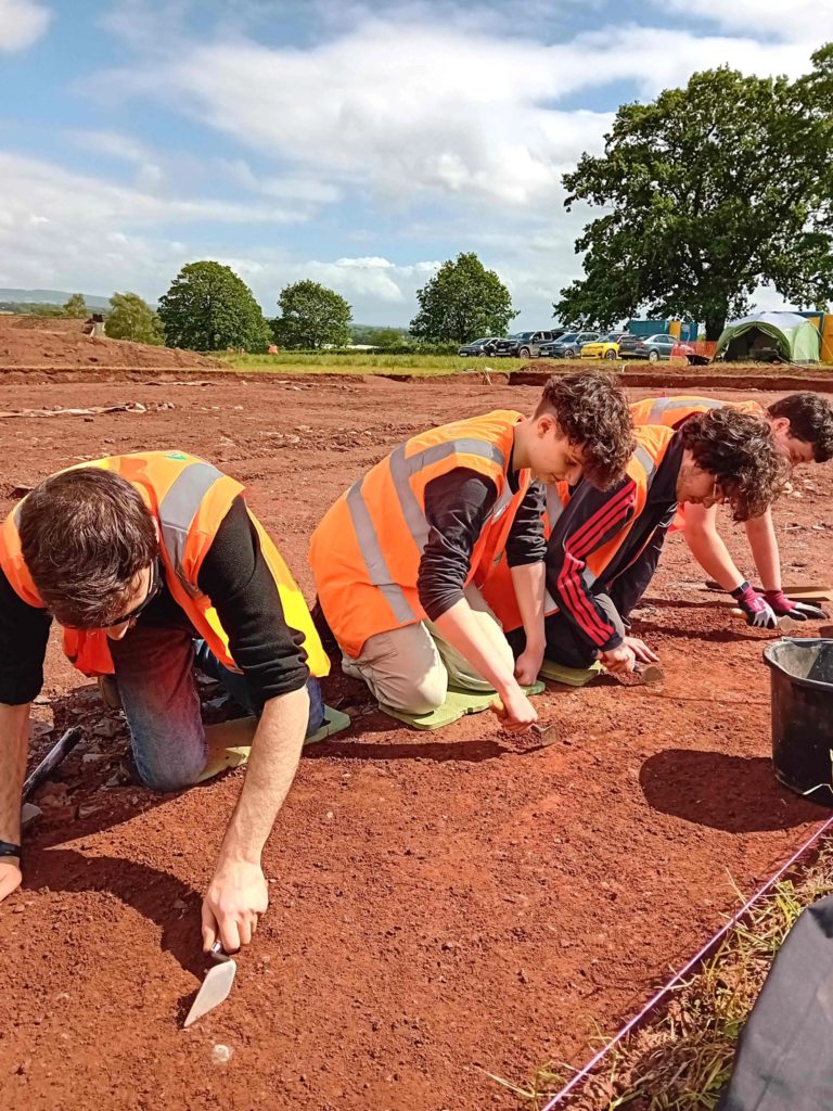 Four students, in high-vis vests, are trowelling at the end of a large excavation area. The soil is bright orange.