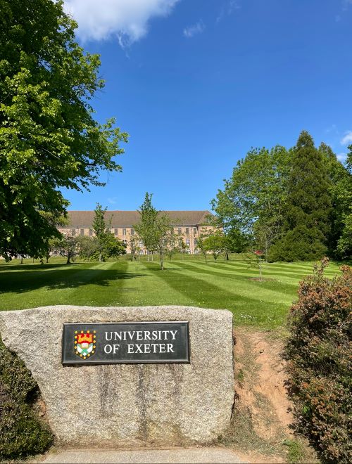 Stone University of Exeter sign in front of a grass lawn and a University building