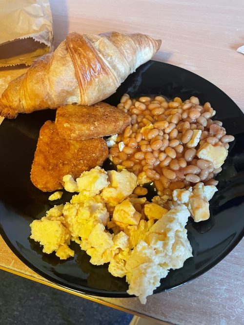 Baked beans, scrambles eggs, hash browns and a croissant on a black plate