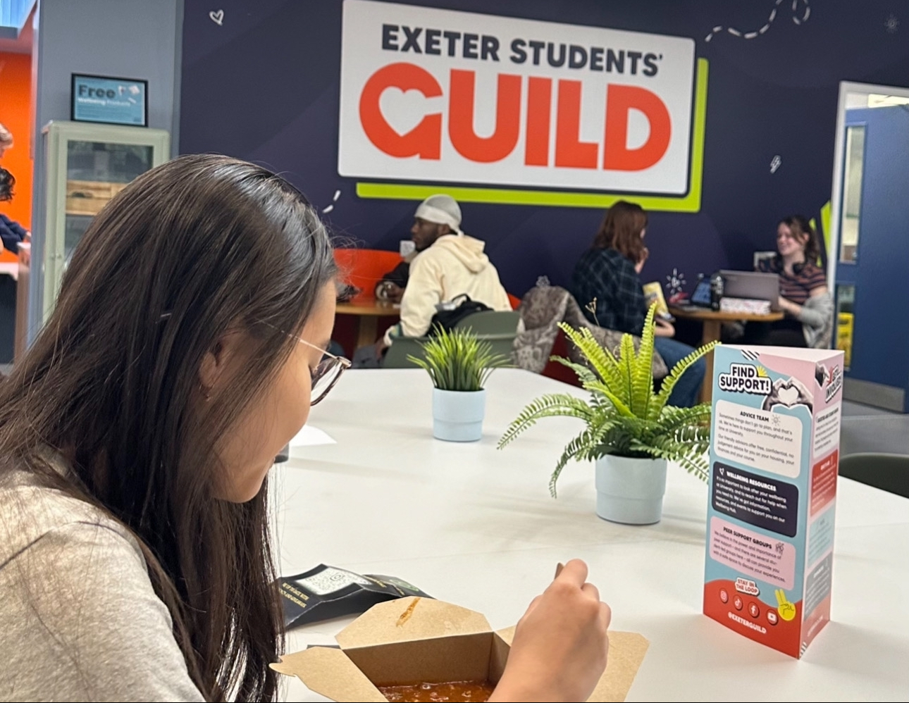 A student eats at a table in the Exeter Students' Guild space, with others chatting in the background.