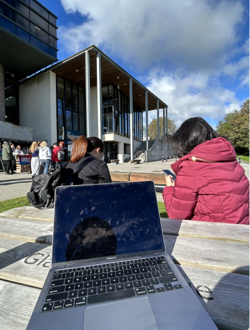 A laptop open on a wooden picnic bench in front of a large, modern building (The Exchange building)