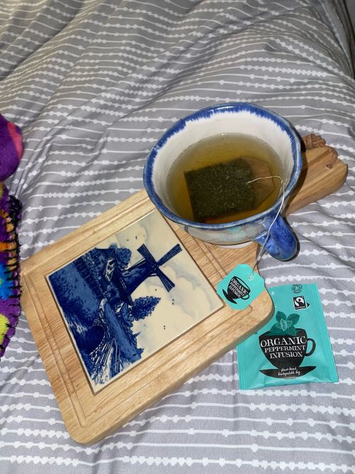 A blue and white china cup of peppermint tea resting on a wooden platter on top of a blue and white striped duvet