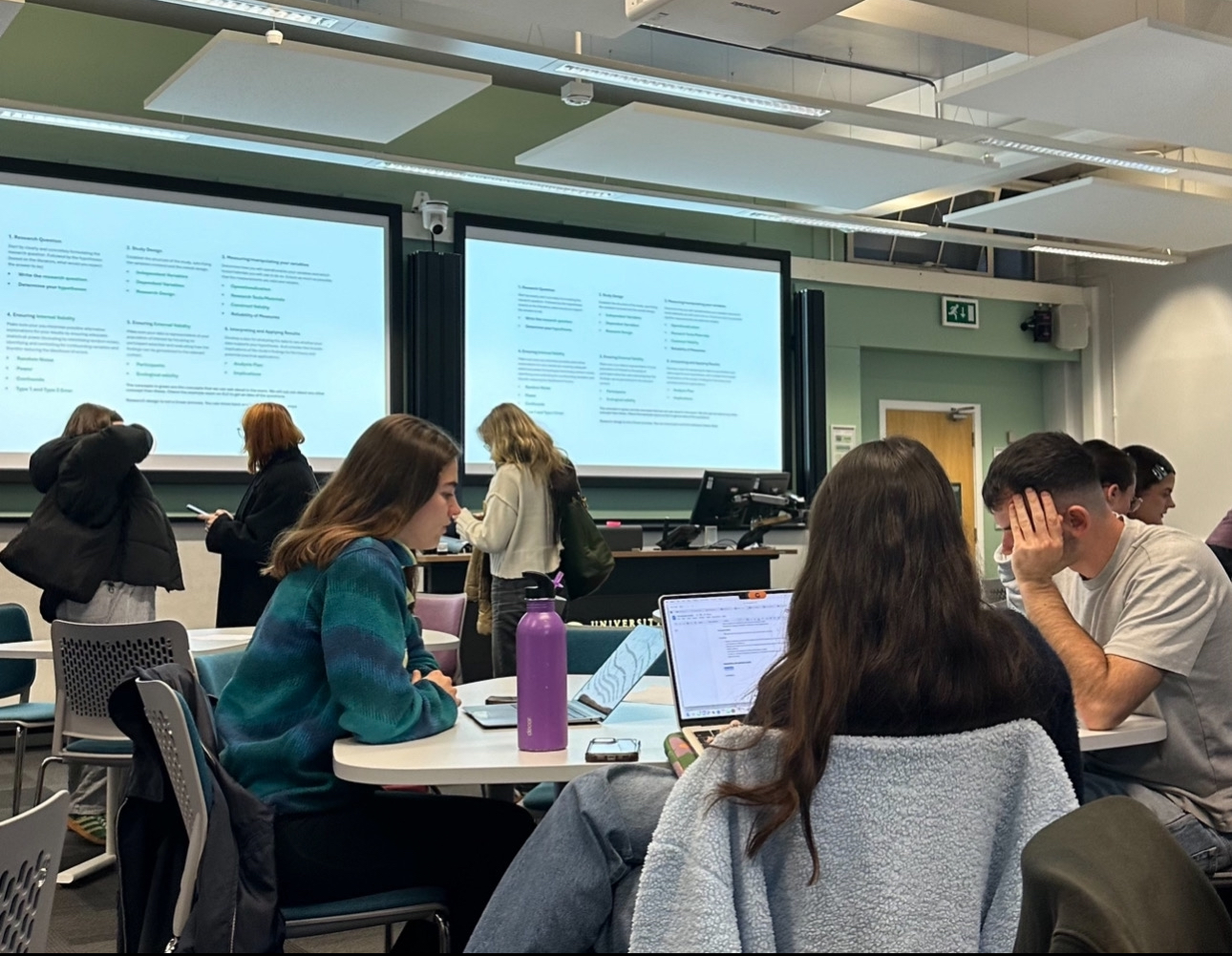 Students in a classroom work on laptops and notes during a statistics practical, with lecture slides projected on the screens at the front.