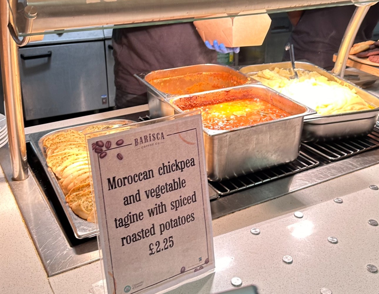 A food counter featuring Moroccan chickpea and vegetable tagine with spiced roasted potatoes, alongside garlic bread and mashed potatoes in metal trays.