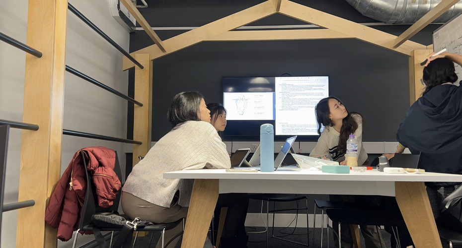 Four students sat at a table full of study materials working together on a large whiteboard