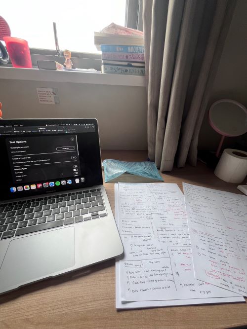 A wooden desk under a window with a laptop and handwritten notes on it.