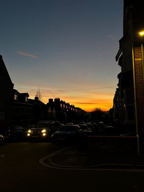 A colourful sunset over a skyline of rooftops and trees along a dark street, with one car's bright headlights pointing towards the camera