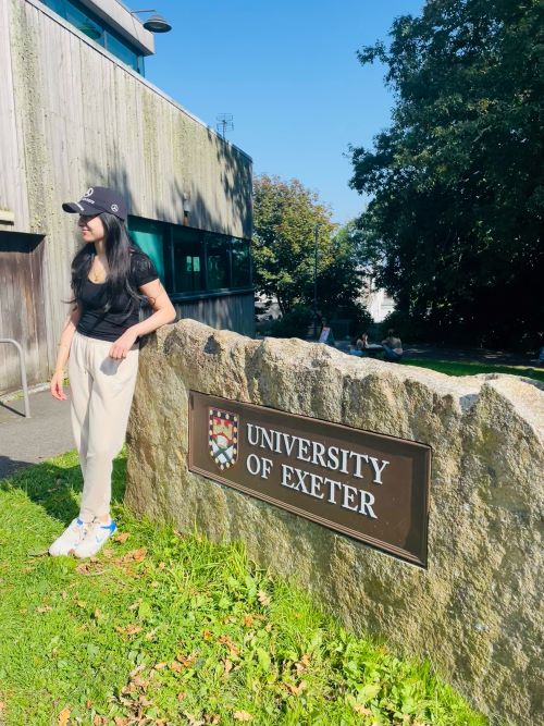 Female student standing on grass on a sunny day leaning against a waist-high rock with a metal 'University of Exeter' sign on it