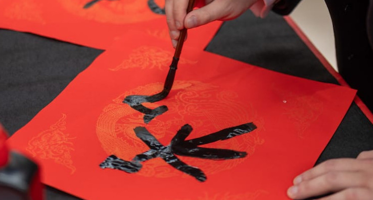 A close-up of a person practicing Chinese calligraphy on a red paper with golden patterns. The individual is using a traditional brush to write black Chinese characters, with another decorated red paper in the background.