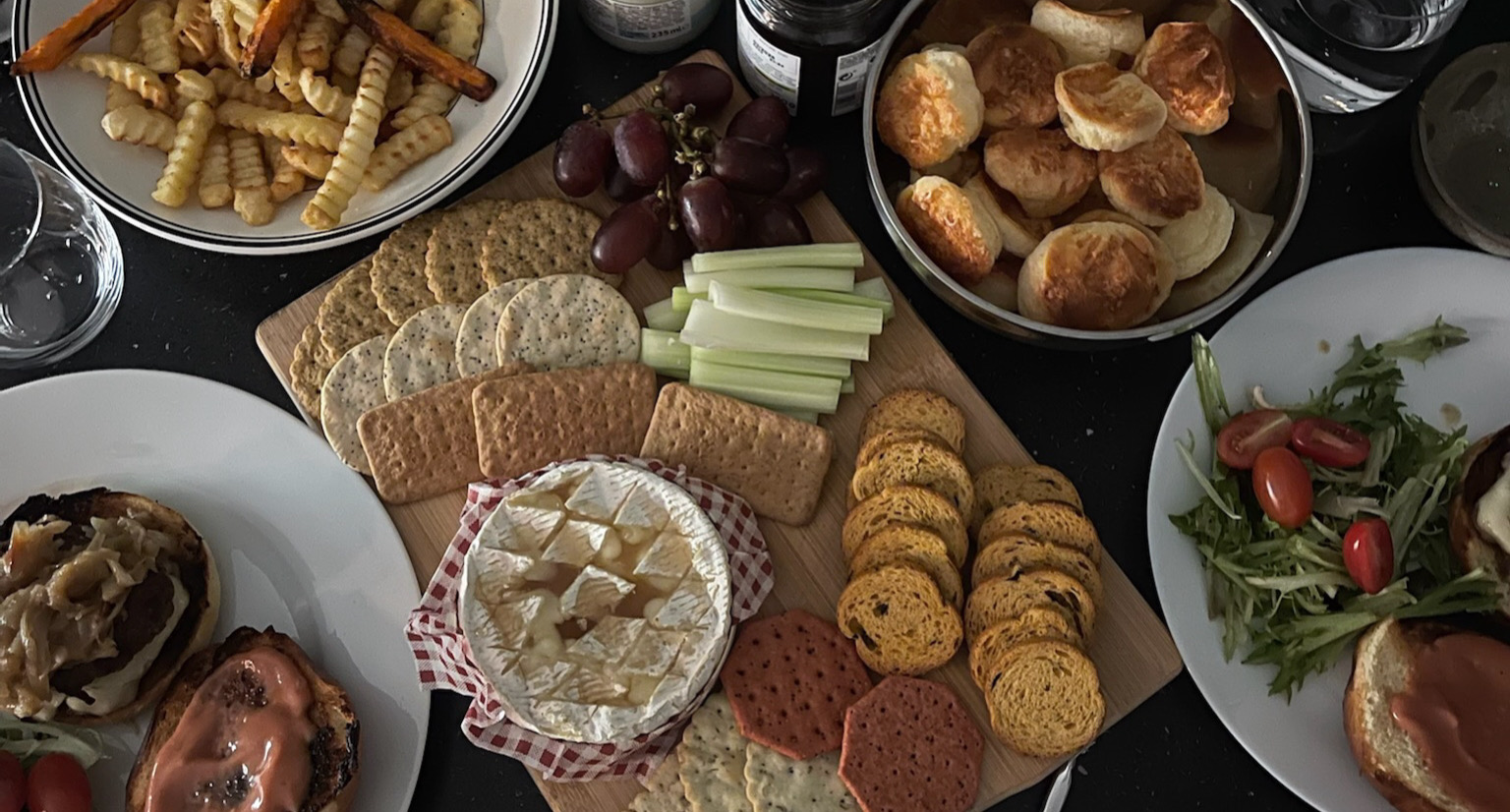 A homemade spread featuring a charcuterie board with baked camembert, assorted crackers, celery sticks, grapes, and cheese scones. Surrounding dishes include burgers, fries, salad, and condiments on a dark tabletop.