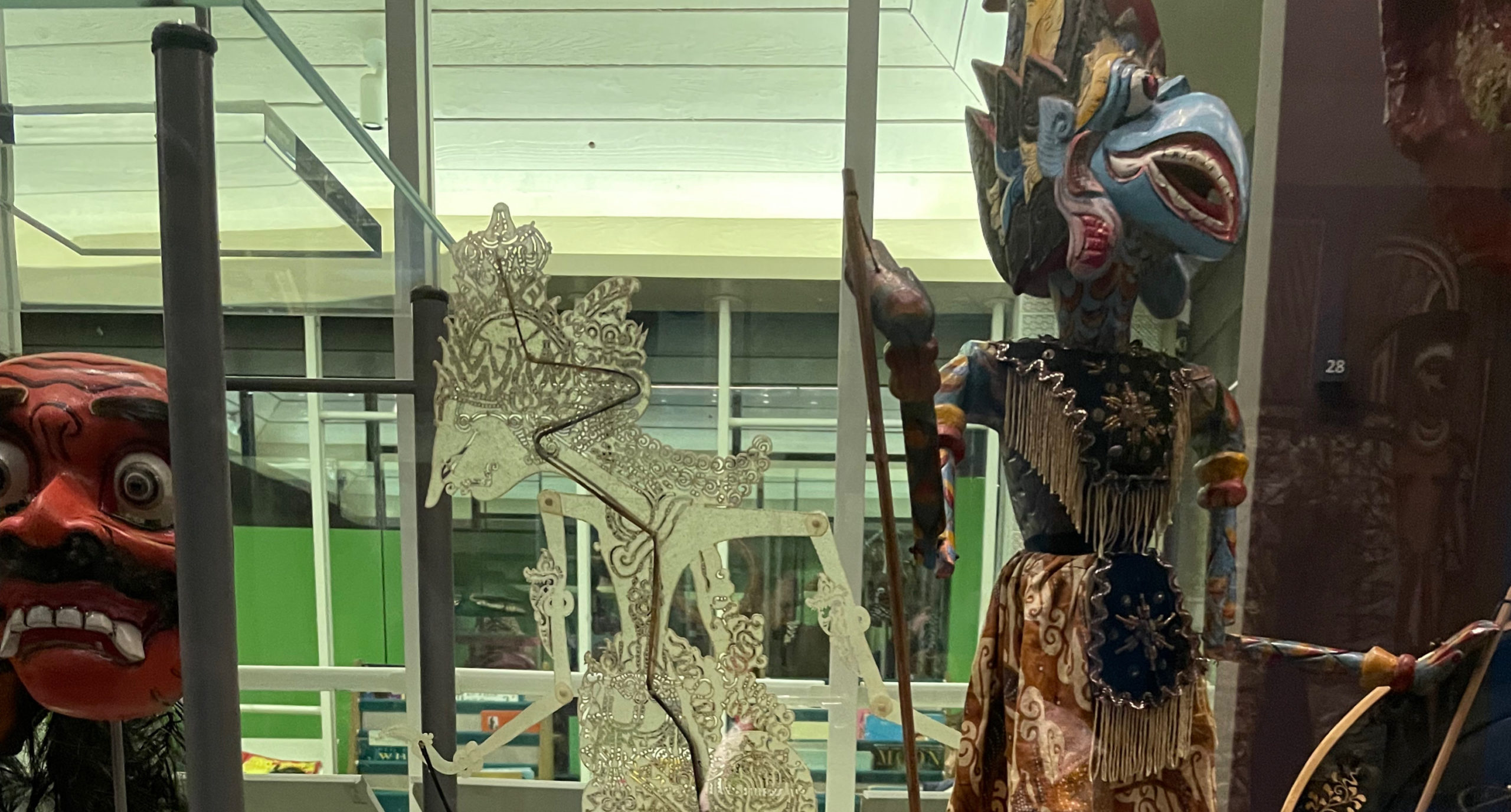 A museum display featuring traditional Indonesian puppets and masks. A wayang kulit (shadow puppet) with intricate carvings stands in the center, alongside a colorful wayang golek puppet dressed in batik fabric. To the left, a red-faced mask with exaggerated features is visible. The display is enclosed in glass with reflections of the surroundings.