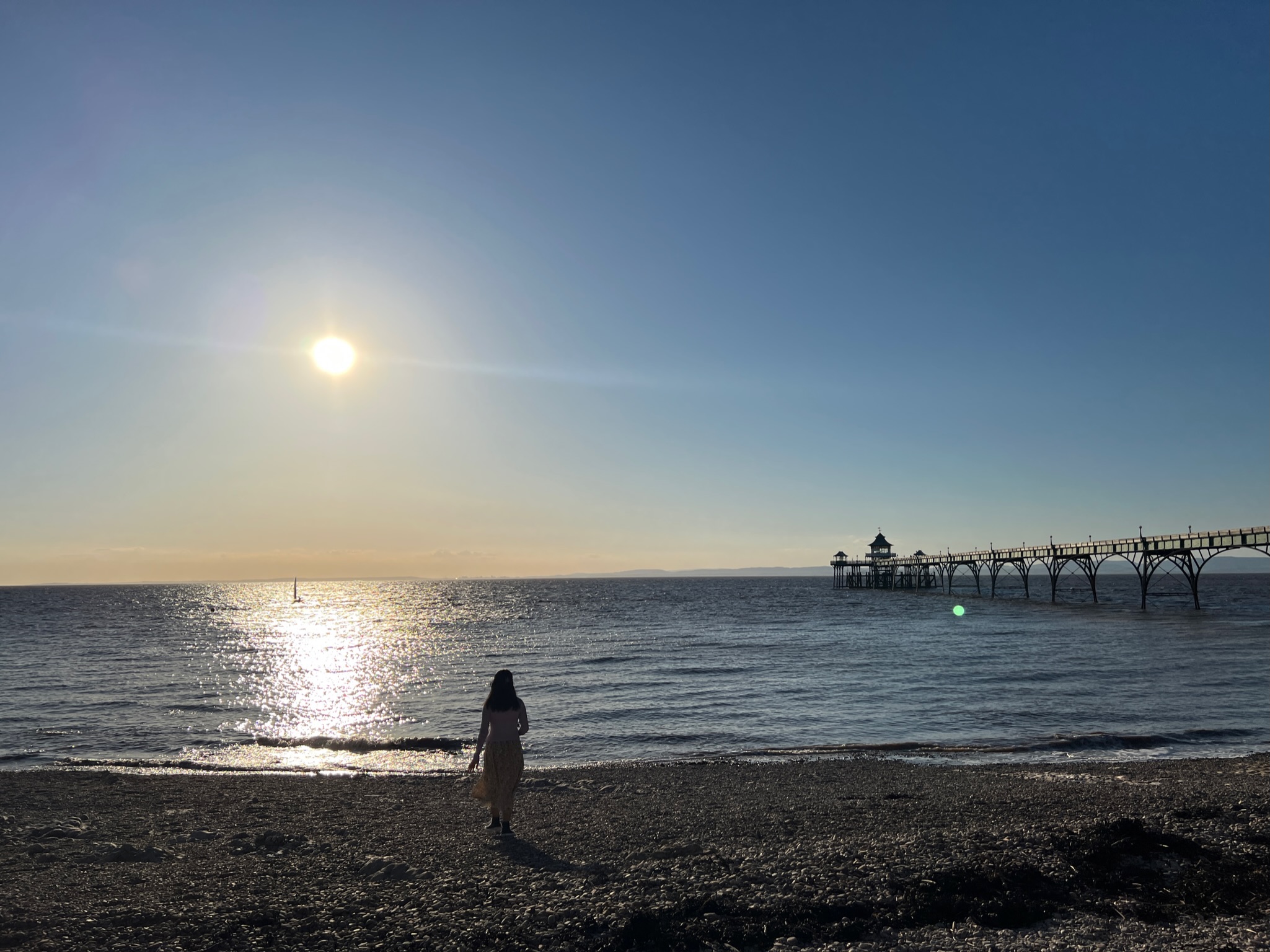 A person walking along the pebbled shore at Clevedon Beach during sunset. The sun reflects off the calm ocean, with a sailboat in the distance and a historic pier stretching into the water.