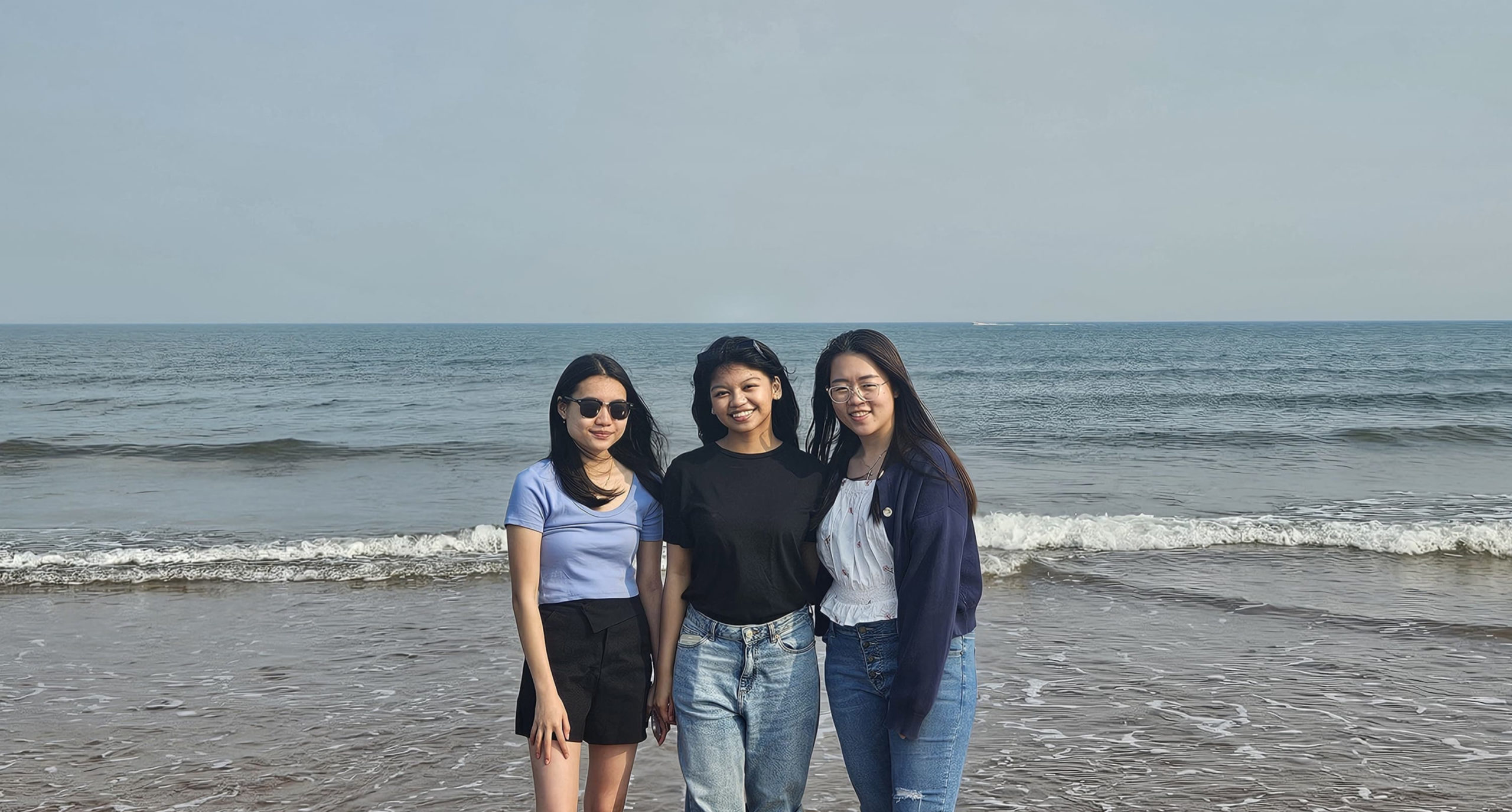 Three friends posing on the beach with the ocean and waves behind them. They are dressed in casual outfits, and enjoying the sunny day.