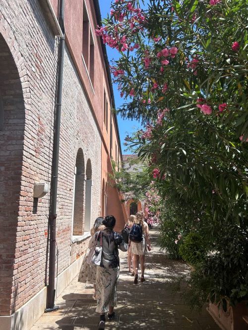 Female students walking between a brick wall and a tall bush covered in bright pink flowers