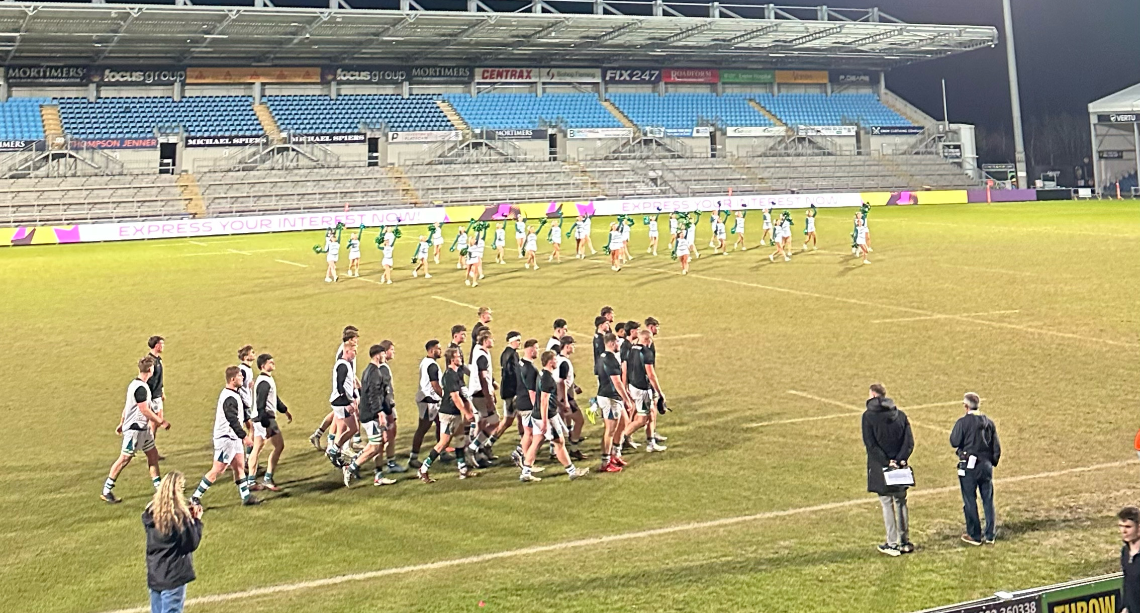 A rugby team walking across the field at night, with cheerleaders performing in the background. The stadium lights are bright, and the stands have some spectators watching.