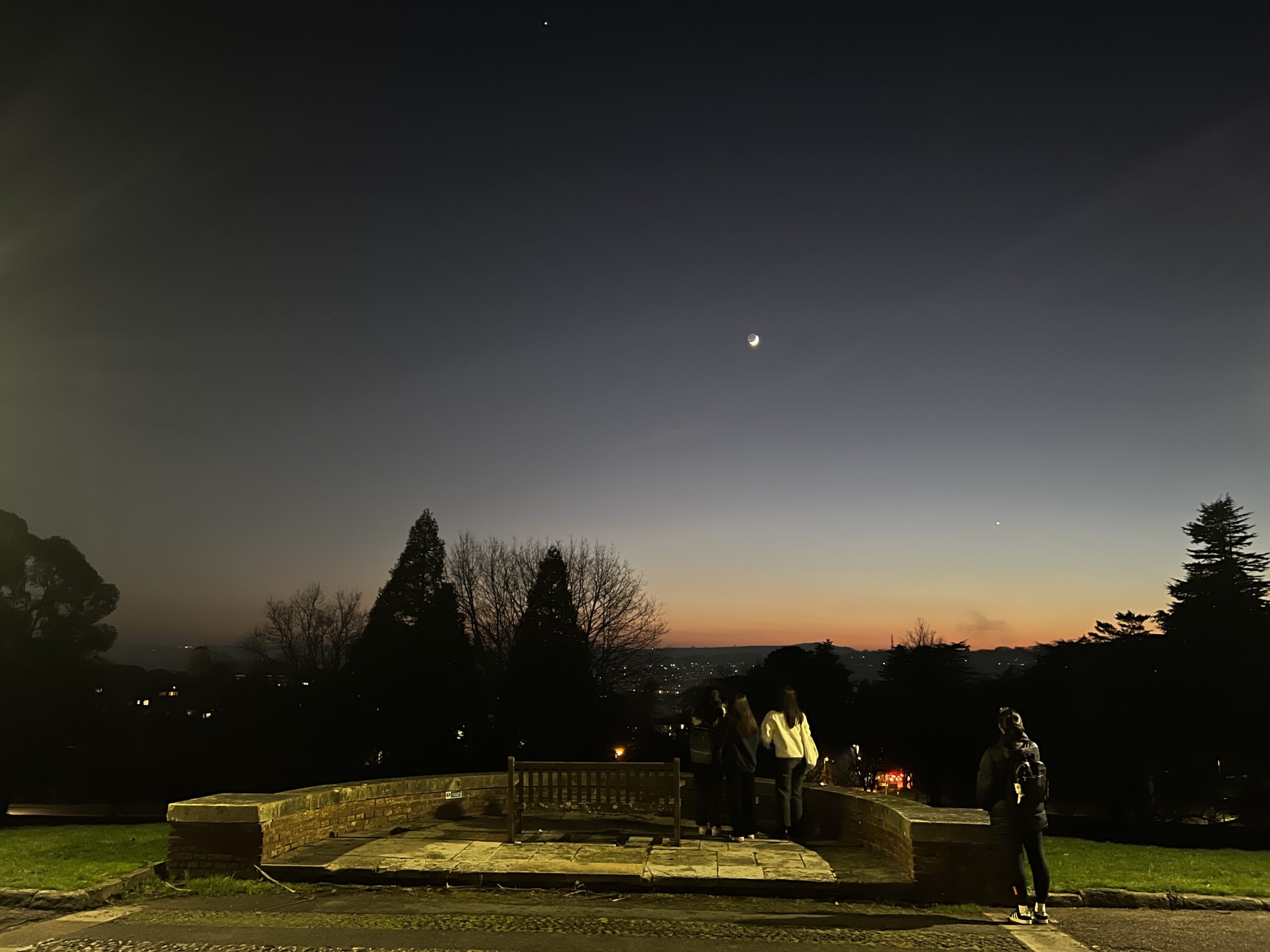 A sunset view from outside Washington Singer in Exeter. The sky fades from deep blue to warm orange, with a crescent moon shining. A few people are gathered by a stone bench, taking in the city lights, while someone with a backpack stands nearby.