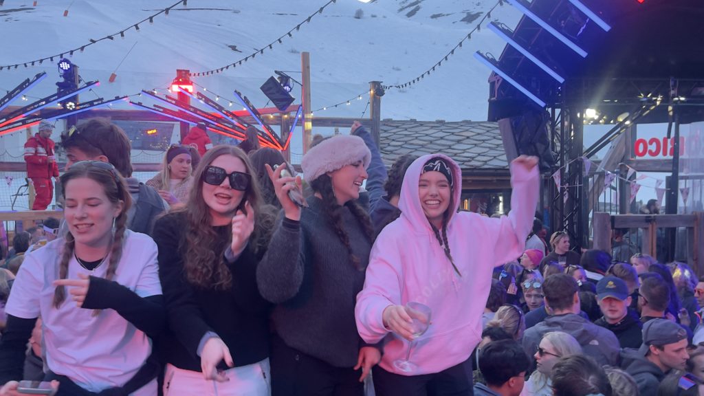 Four girls dancing on table tops at an apes bar in France with a crowd of people and mountains in the background.