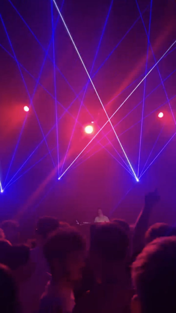 Lasers in white, purple, and pink with a crowd waving their hands in the air and DJ in the centre