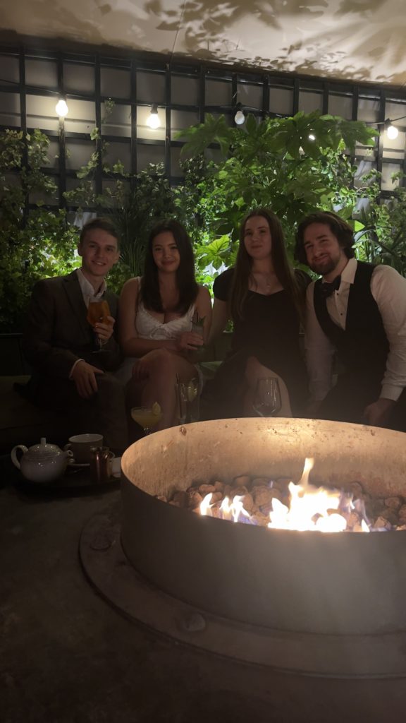 Four young people sitting around a fire pit on a balcony holding drinks with a live wall behind them.