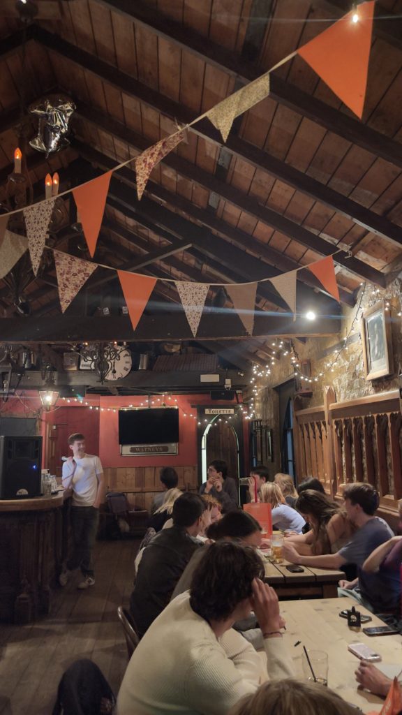 Loft style pub with people seated at tables for a quiz. One stands to the side with a microphone. Colourful orange patterned bunting and fairy lights decorate the room
