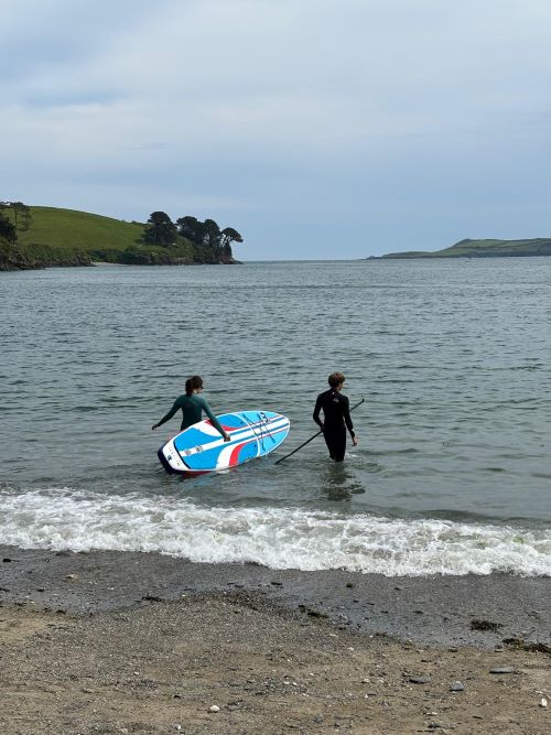 Two students in the shallow water in the sea, one carrying a blue paddleboard