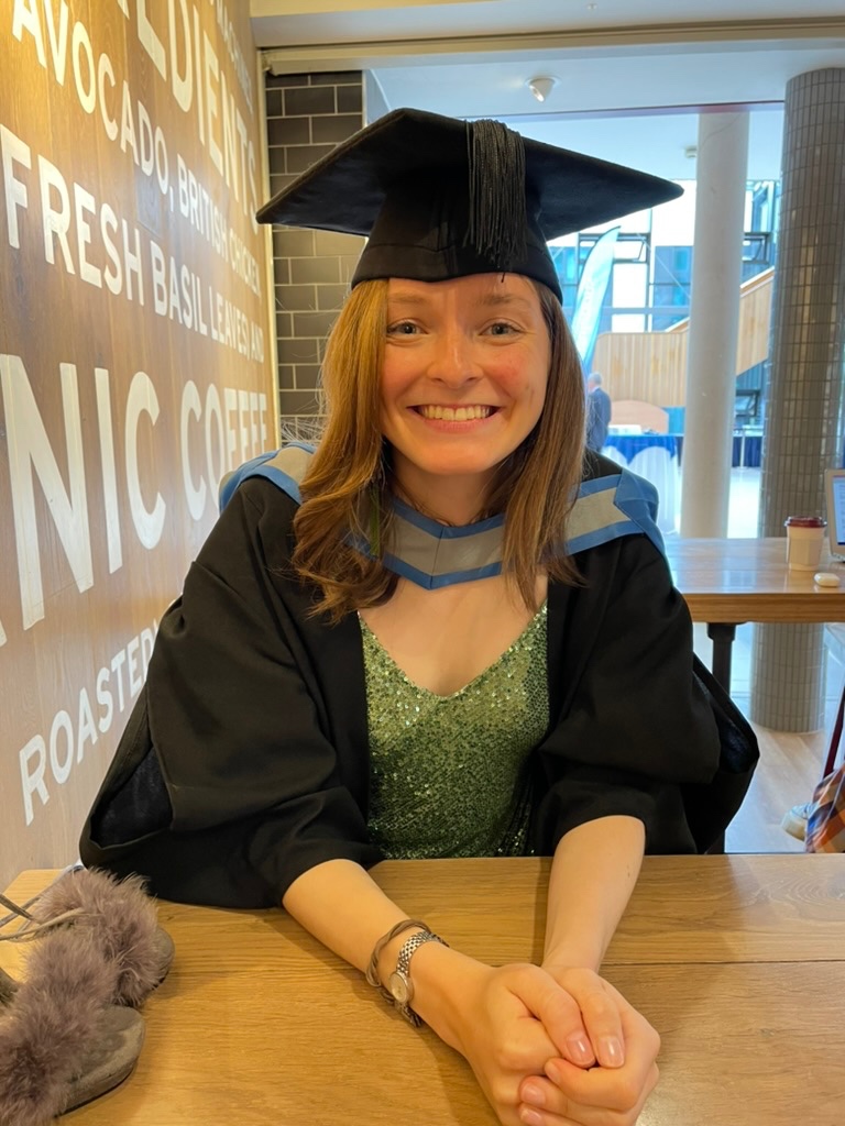 Photo of a female graduate wearing their cap and gown sat in Pret a Manger in the Streatham Campus Forum building
