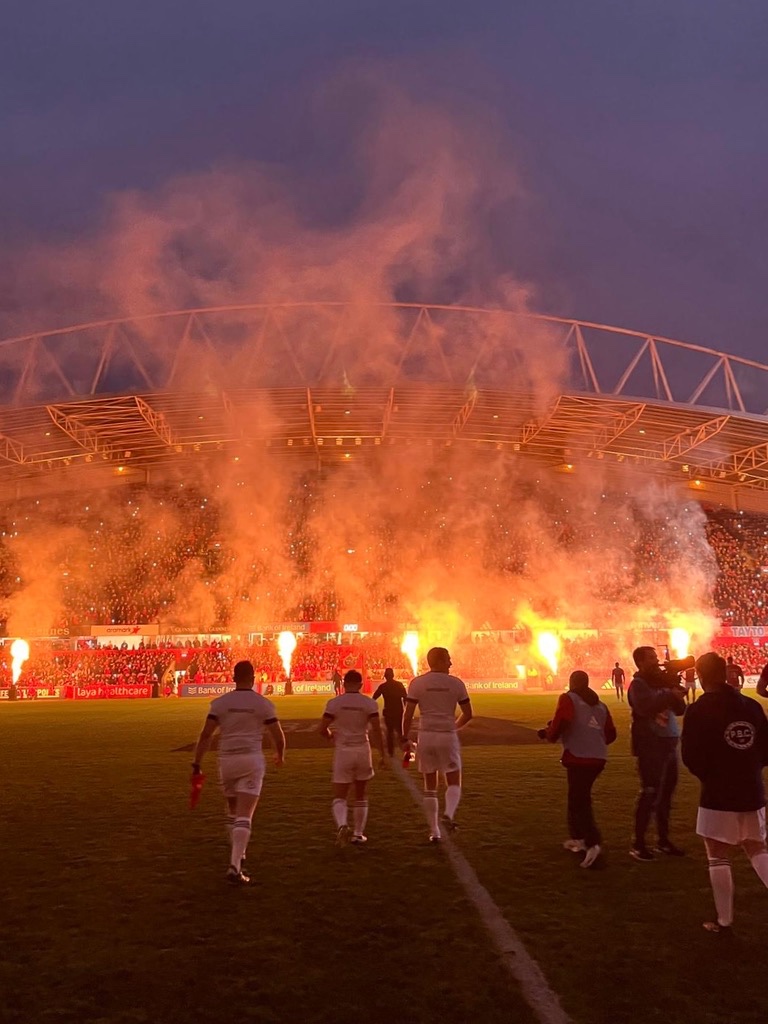 Photo of rugby players running out onto a pitch at night, with flares going off on the sidelines