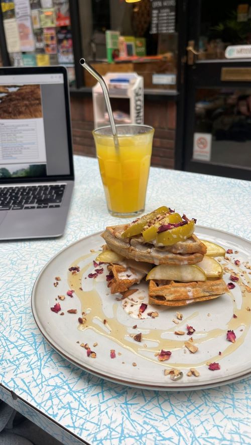 A plate of fruit-topped waffles and a glass of orange juice on a table with a laptop