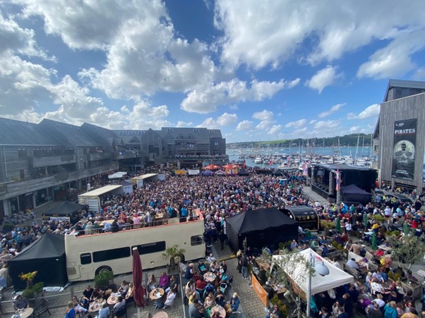 Crowded piazza with a large concert stage next to a harbour full of boats