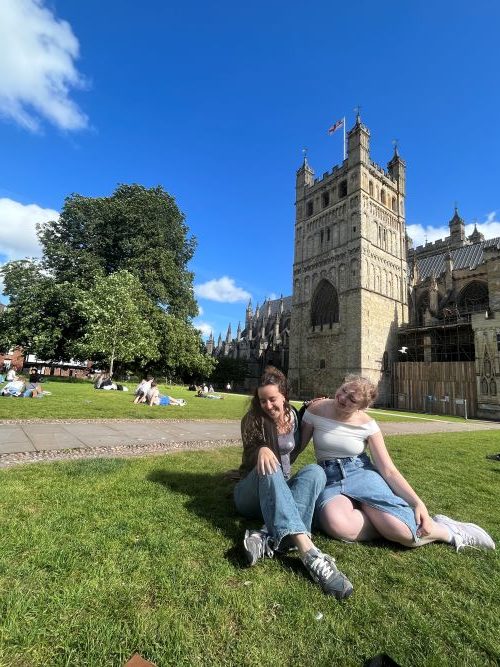 Two female students smiling with an arm around one another sat on the grass in front of Exeter cathedral on a sunny day