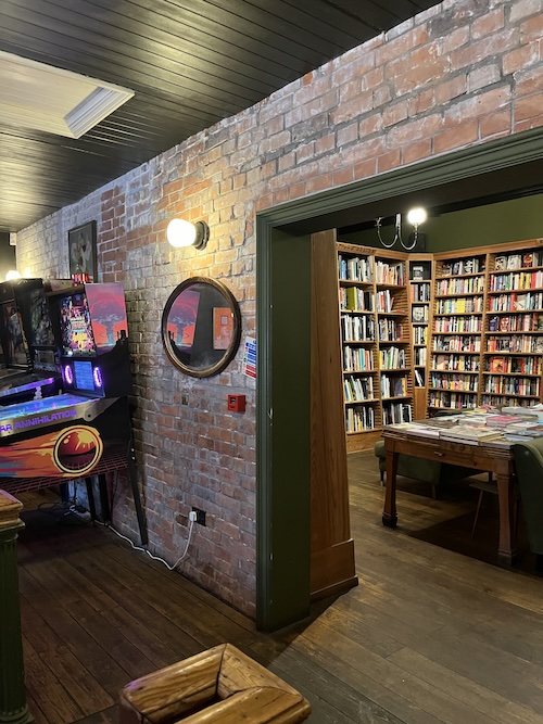 The inside of a pub with retro arcade machines on one side and bookshelves on the other side through a brick doorway
