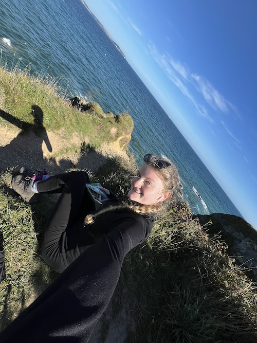 A selfie of me sitting on a grass cliff, with a background of blue sea and blue sky, holding a pasta salad and smiling