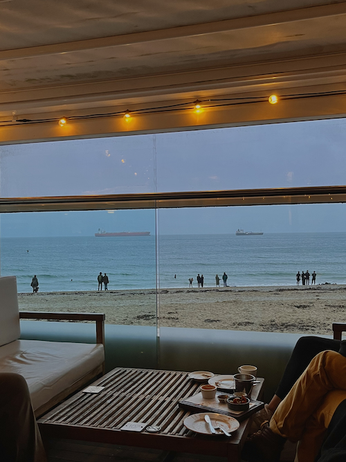 A cosy cafe scene with fairy lights, comfy chairs, mugs and plates, and a beach and sea view out of the window