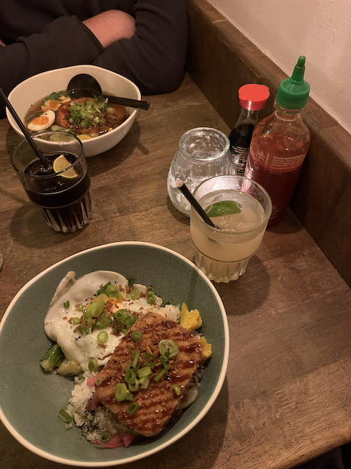 A candlelit table with two soft drinks, bowls of ramen and donburi with garnish, and sauces