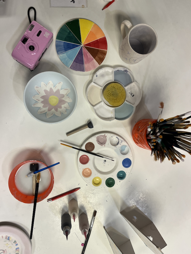 A table with paints, paintbrushes, colour wheel, film camera, and half painted mug and bowl