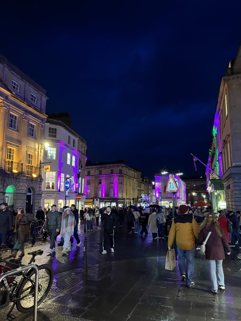 Crowds on a street in Bath city centre, showing a dark evening sky and purple lighting on nearby buildings.