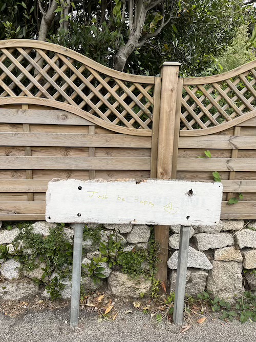 A white disused sign on a street with the words 'just be happy' written in gold pen, with a picture of a sail boat 