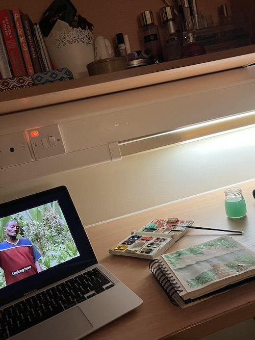 A cosy desk in student accommodation with a documentary on a laptop next to a watercolour set, paintbrush and notebook with a half completed painting 