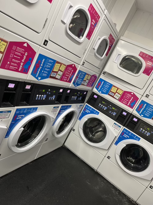A photo of a laundry room in student accommodation, with multiple washing machines next to each other, and dryers on top in a separate row. There are lots of clear stickers and instructions on how to use the machines