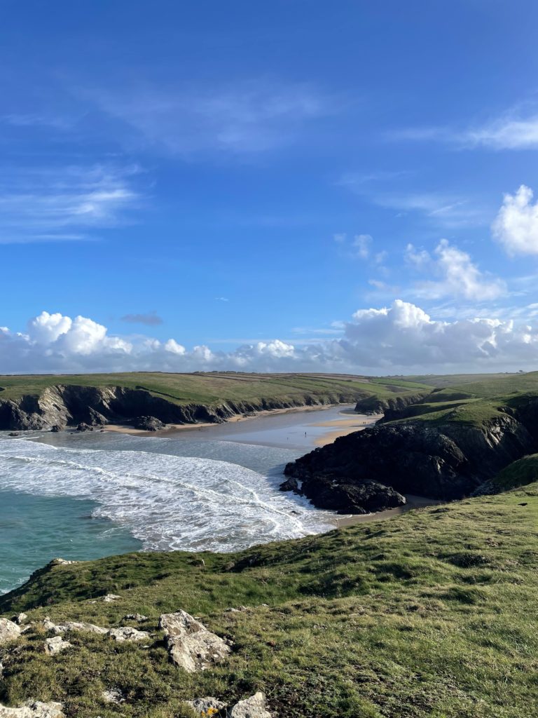 A beach cove with two surrounding headlands. Bright blue sky and sea with flat green fields with cliffs dipping down into the sea.