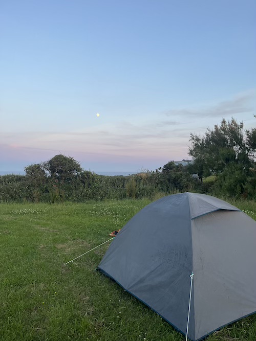 A small grey tent on a lush green field. There is a hedge and the horizon of the sea in the background. The sky is blue and pink with a visible moon.