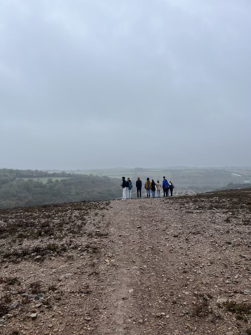 A group of students standing on a barren landscape, with brown stones and vegetation. It's an atmosphere foggy, overcast day. There are green fields in the background which contrast the landscape of the so-called Cornish Mars
