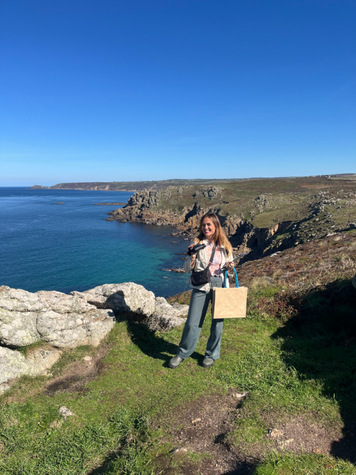 Tatiana holding binoculars and a bag and standing on a headland with the coastline and sea beyond.
