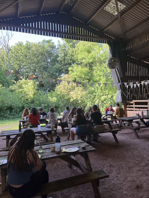 Students sitting at picnic benches in a farm barn structure, decorated with bunting, fairy lights and a disco ball. Golden sunlight on trees behind the barn.