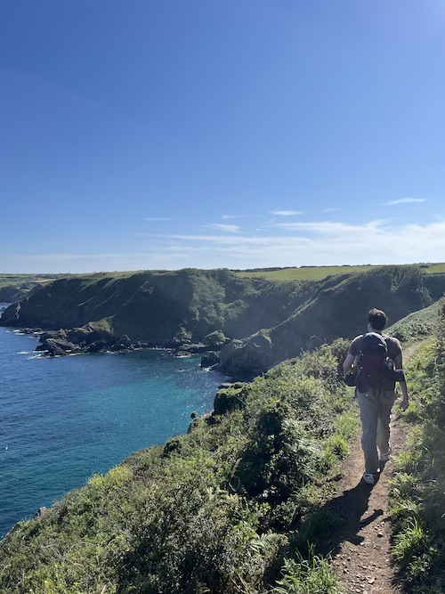 A man walking on the coast path with a hiking rucksack on. There are blue skies, bright green fields and cliffs, and vibrant turquoise sea below the path.