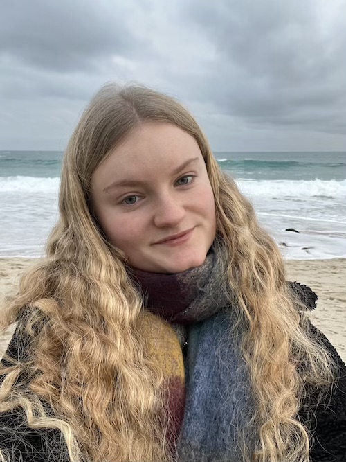 A selfie of me, a woman with blonde wavy hair, wrapped up in a coat and scarf, smiling. In the background is a beach, turquoise sea and a cloudy sky.