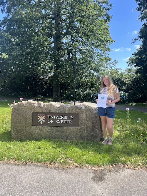 A woman with blonde hair, shorts and t-shirt, holding up a dissertation in front of a rock with the University of Exeter sign. It's a sunny day with bright green grass and blue sky.